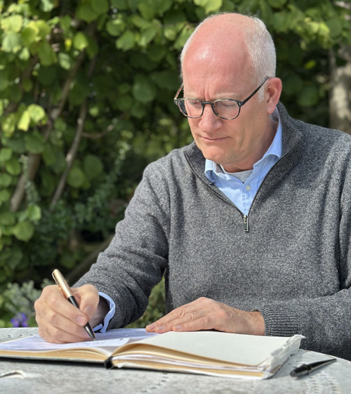 Portrait of Piers Clemett writing at a table in the garden